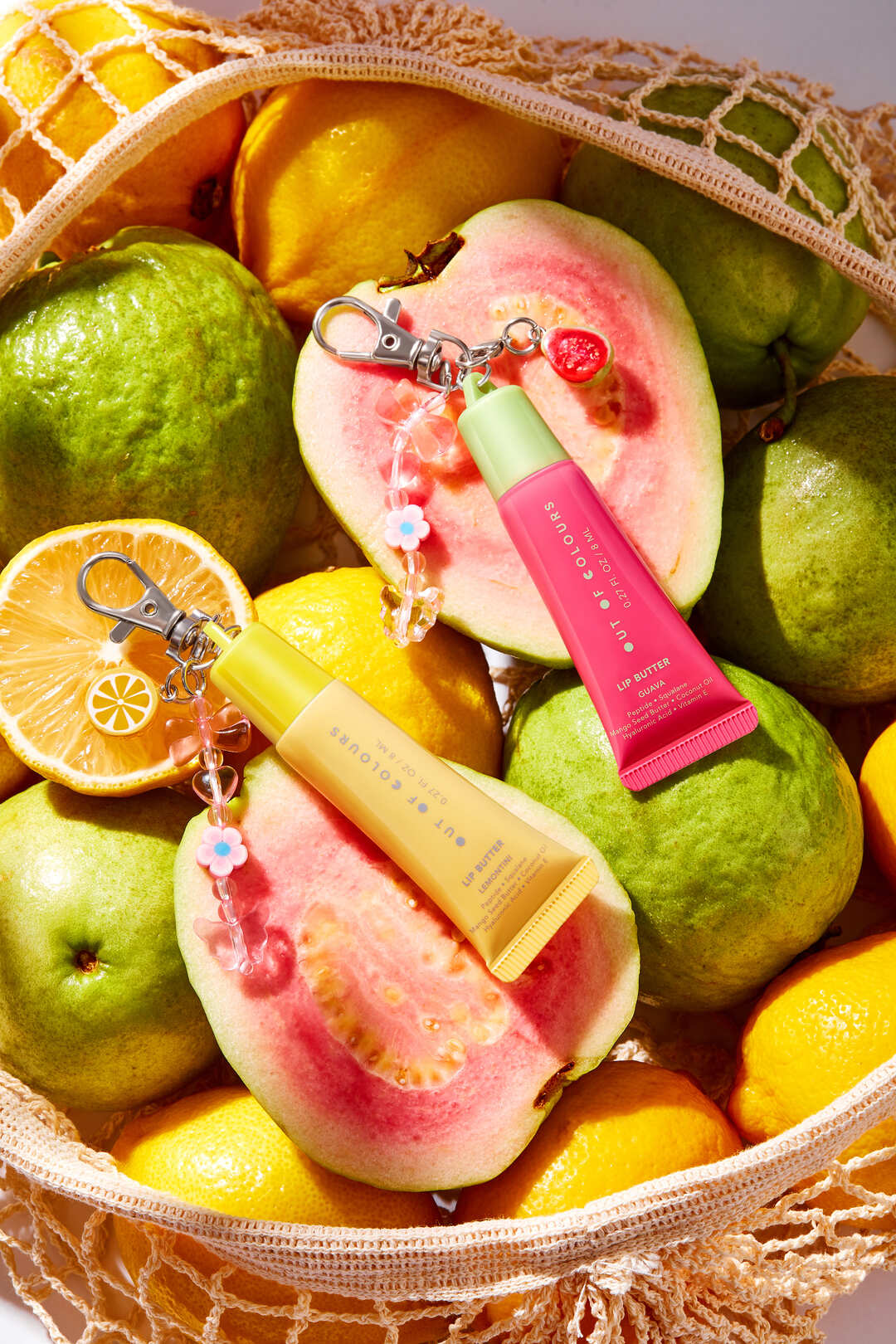 Out of Colours Lip Butter Balms in Guava and Lemontini with charm keychains, surrounded by fresh guavas and lemons