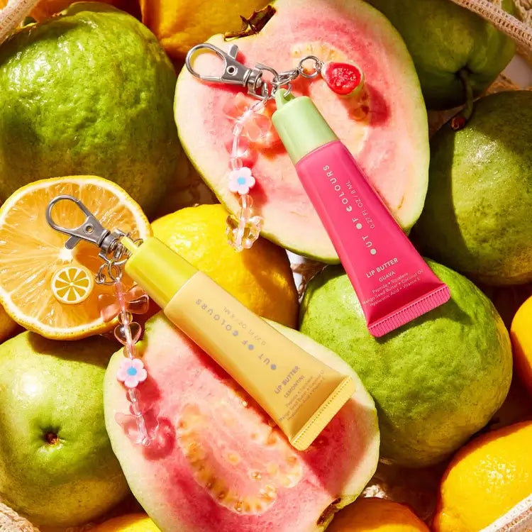 Two lip balm tubes with floral keychain charms are arranged on a bed of guava fruits, oranges, and a sliced guava half. The lip balm tubes are one bright pink with a light green cap and one pale yellow with a light blue cap.